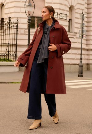 Femme marchant dans la rue de la ville portant un long manteau couleur rouille, un pantalon large foncé, des bottes beiges et des boucles d'oreilles en or.