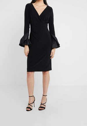 Femme portant une robe portefeuille noire mi-genou avec un décolleté en V et des manches évasées bordées de satin, associée à des sandales à talons hauts noires à lanières.