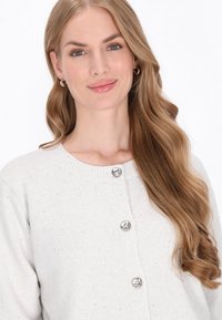 Cardigan gris clair en tissu tricoté doux, avec un col côtelé et trois boutons argentés à l'avant. Texture subtile sur l'ensemble.