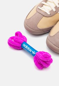 Adidas-märkt leksak i ett fluffigt rosa material, formad som en hantel, placerad bredvid bruna mockaskor med beige detaljer och vita snören.