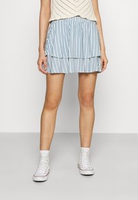 Donna che indossa una minigonna a strisce blu e bianche a strati, un top lavorato a maglia bianco, calze bianche e sneakers alte bianche, che si trova in piedi contro uno sfondo neutro.