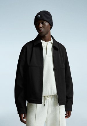 Black zip-up jacket with a textured finish, worn over a light beige polo shirt. The model wears a black ribbed beanie. Neutral background.