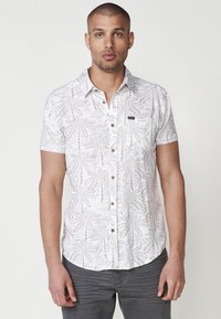 Camisa blanca de manga corta con un patrón de hojas grises, cierre de botones en la parte delantera y un pequeño parche con logo. Tejido suave con un corte relajado.