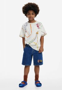 Chemise blanche à manches courtes avec des motifs abstraits colorés, assortie à un short en jean bleu orné d'un écusson décoratif. Le modèle porte des chaussettes marron et des sandales bleues.