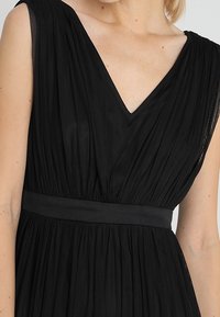 Černé šaty s výstřihem do V, skládacím tylovým materiálem, širokými ramínky a dobře padnoucím pasem. Jemná textura s vrstveným designem.