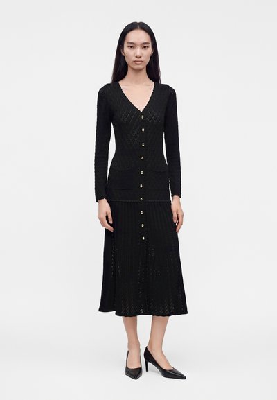Femme portant une robe en maille noire à manches longues avec des boutons dorés sur le devant et des talons noirs à bout pointu, debout devant un fond uni.