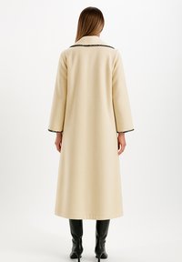 Femme portant un long manteau beige avec des finitions noires sur le col et les poignets, associé à des bottes noires à talons montantes jusqu'aux genoux, se tenant de dos.