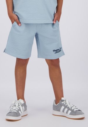 Kind staat met handen in de zakken, draagt lichtblauwe gestructureerde korte broek en grijze sneakers met witte veters en strepen op een witte achtergrond.