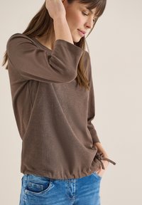 Jeune femme ajustant un haut marron à manches longues avec un détail noué à l'ourlet, portant un jean bleu, sur un fond beige uni.