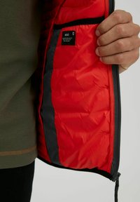 Interior de una chaqueta acolchada roja, con textura de acolchado, que presenta una cremallera negra y una etiqueta. Mano sosteniendo la chaqueta contra un suéter verde.