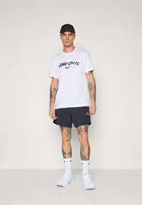 Vit t-shirt med marinblå "Just Do It" text, svarta träningsshorts, vita sneakers och en marinblå keps. Tatueringar syns på armar och ben.