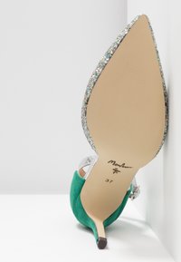 Grön högklackad sko med en silvrig glittrande sula, vinklad mot en vit bakgrund, som visar sitt märke och storlek under hälen.