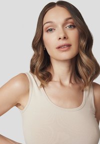 Beżowy top na ramiączkach z okrągłym dekoltem i prążkowaną fakturą. Modelka ma falujące, sięgające ramion brązowe włosy oraz minimalny makijaż.