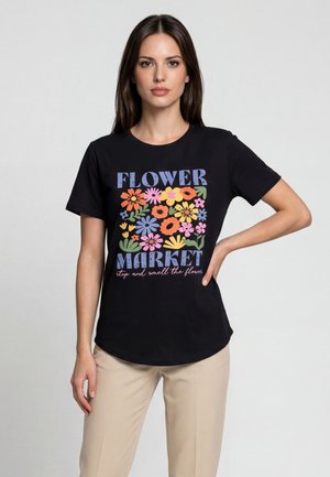 Donna che indossa una t-shirt nera con grafica floreale colorata e testo "Flower Market stop and smell the flower," abbinata a pantaloni beige.