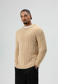 Beige kabelgebreide trui met een zachte, textuurrijke afwerking en een ronde hals. Model heeft handen in de zakken, gecombineerd met zwarte broek.