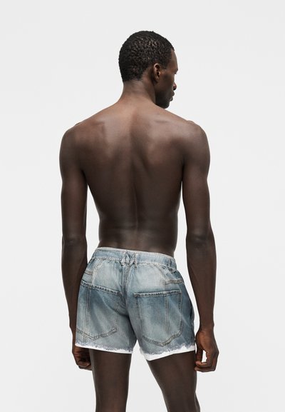 Άντρας με σκουρόχρωμο δέρμα, κοντά σγουρά μαλλιά, χωρίς πουκάμισο, φοράει ανοιχτόχρωμα τζιν σορτς, στέκεται με την πλάτη προς τα εμπρός μπροστά σε απλό λευκό φόντο.