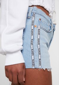 Shorts de mezclilla azul claro con dobladillo deshilachado, con cinta de marca en blanco y negro a lo largo de las costuras laterales. Llevado con una camiseta blanca de manga larga.