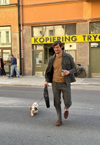 Man in een gestreepte grijze blazer over een bruine trui, grijze broek, die een zwarte tas en een koffie draagt, terwijl hij een klein wit hondje met bruine vlekken uitlaat.