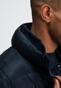 Marineblauer Puffer-Jacke mit hohem Kragen, gestepptem Design und strukturiertem Innenkragen. Mit einem Frontreißverschluss und Knopfdetails.