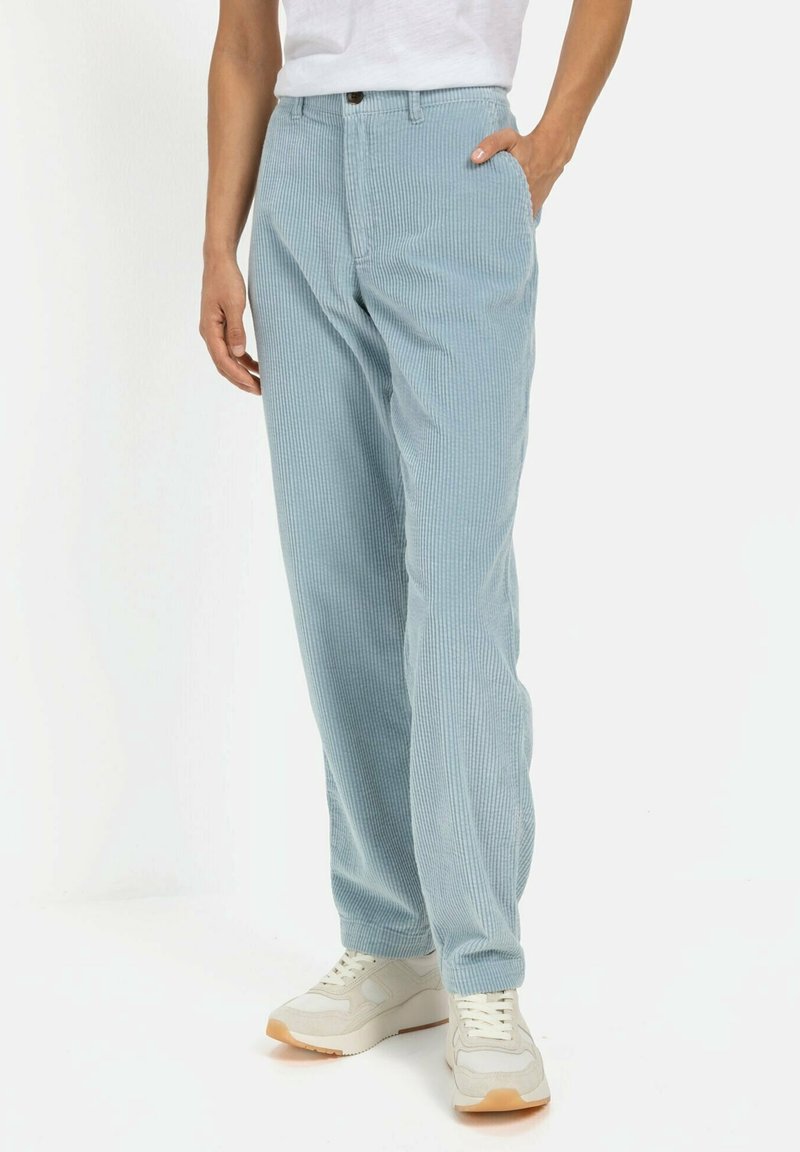 Personne portant un pantalon en velours côtelé bleu clair et des baskets blanches, debout avec une main dans la poche devant un fond uni.