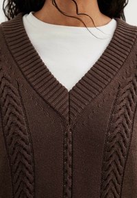 Brauner gestrickter Pullover mit V-Ausschnitt, der strukturierte Zopfmuster an den Seiten und Rippabschlüsse aufweist. Getragen über einem weißen Hemd.