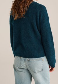 Petrolejově zelený chlupatý svetr s volným střihem a žebrovaným výstřihem, nošený s světle modrými džínami s kapsami na zádech a světlým efektem praní.