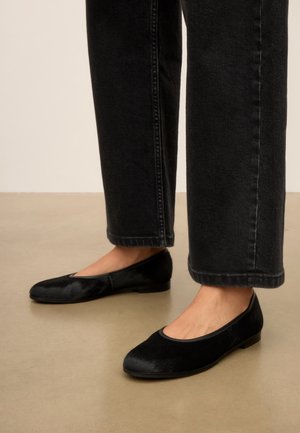 Persoon met zwarte rechtvallende broek en zwarte fluwelen ballerina's staand op een beige vloer.