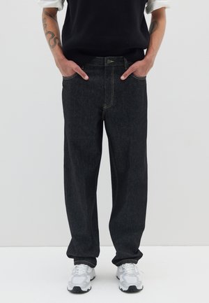 Persona con jeans negros de corte holgado, zapatillas blancas y un suéter negro sin mangas sobre una camisa blanca, con las manos en los bolsillos, de pie contra un fondo blanco.