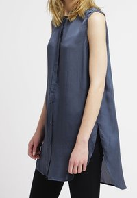 Ärmelloses, dunkelblaues Hemd mit Knopfleiste aus strukturierter Stoff, mit asymmetrischem Saum und Seitenschlitzen, getragen über schwarzen Hosen.