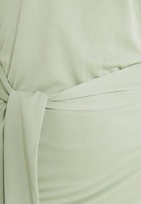 Jasnozielona bluzka z wiązanym paskiem, o gładkiej fakturze i luźnym kroju, podkreślająca opadającą sylwetkę.