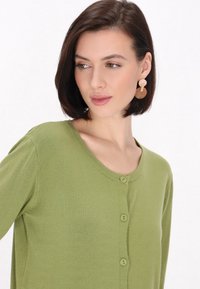 Cardigan verde realizzato in materiale morbido con scollatura rotonda e cinque bottoni davanti. Il modello indossa grandi orecchini circolari color crema.