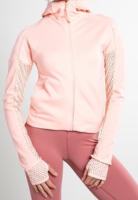 Light pink zip-up athletic jacket with perforated sleeves and thumb holes, worn with mauve fitted leggings on a standing person.