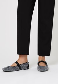 Pantofole grigie pelose con una strap nera e un accento lucido. Il design presenta una punta rotonda e una texture morbida, abbinate a pantaloni neri.