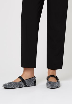 Pantofole grigie pelose con una strap nera e un accento lucido. Il design presenta una punta rotonda e una texture morbida, abbinate a pantaloni neri.