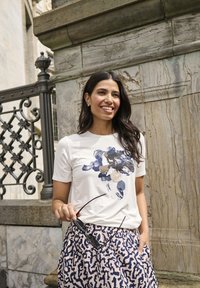Weißes T-Shirt mit marineblauer Blumengestaltung, kombiniert mit einem gemusterten rosa Rock mit dunkelblauen Akzenten. Das Model hält eine Sonnenbrille und steht vor einer Steinmauer.