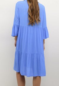 Blå klänning med trekvartslånga ärmar, i lagerdesign och mjukt, texturerat tyg. Har en utsvängd form och rundat fåll.