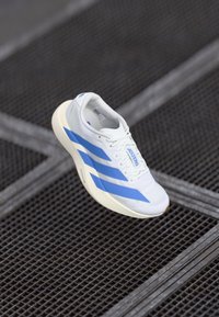 Pantofii sport albi cu dungi albastre, design ușor, superior din plasă, talpă texturată și sistem de șireturi minimalist.