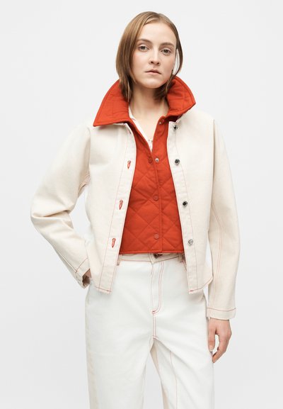 Λευκό τζιν μπουφάν με κόκκινες λεπτομέρειες, φορεμένο πάνω από κόκκινο καπιτονέ γιλέκο. Σημαντικά χαρακτηριστικά περιλαμβάνουν κουμπιά πατιλέτας και εμφανείς ραφές.