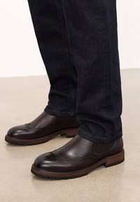 Stivali brogue in pelle marrone con una finitura liscia, caratterizzati da perforazioni decorative, una punta arrotondata e una suola in gomma testurizzata.