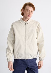 Lys beige jakke med lynlås lukning, elastiske manchetter og et lille logo på venstre bryst. Glat stof, afslappet pasform og krave-design.