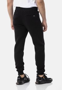 Schwarze Jogginghosen aus weichem Stoff, mit schmalem Schnitt, elastischem Taillenbund und zwei Rückentaschen mit Reißverschlüssen. Kombiniert mit schwarzen Sneakers.