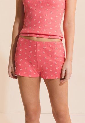 Mujer con top de tirantes de punto waffle rosa y pantalones cortos a juego con un pequeño estampado floral, de pie contra un fondo neutro.