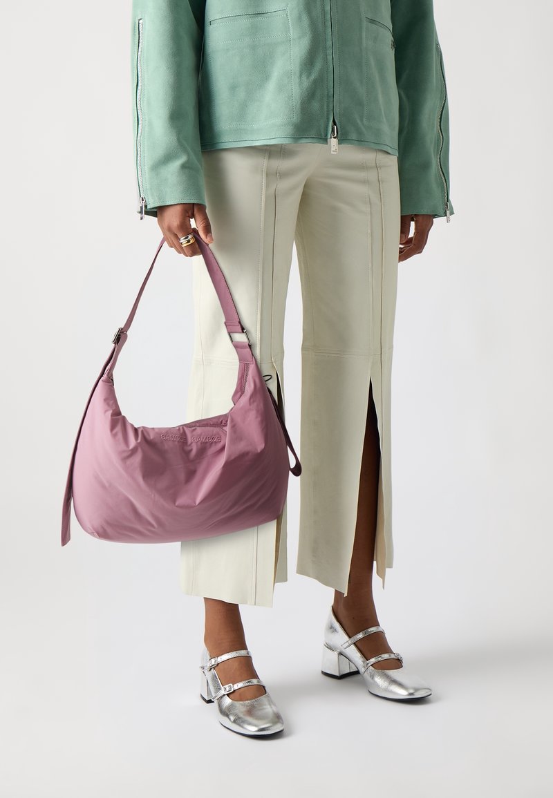 Personne tenant un sac à main mauve, portant un pantalon beige clair avec des fentes à l'avant, des chaussures Mary Jane argentées et une veste zippée verte.