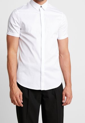 Hombre vistiendo una camisa blanca de manga corta ajustada con cuello puntiagudo y pantalones negros, sobre un fondo liso.