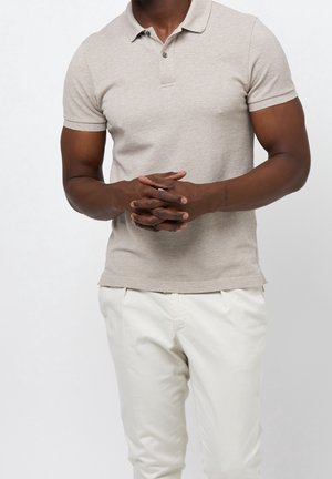 Mężczyzna w beżowej koszulce polo z krótkim rękawem i białych spodniach, stojący z splecionymi dłońmi na tle jednolitej, białej ściany.