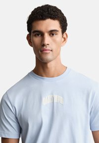 Svetlo modra bombažna majica s kratkimi rokavi, z ukrivljenim vezenim belim logotipom "GANT 1949" na sprednji strani.