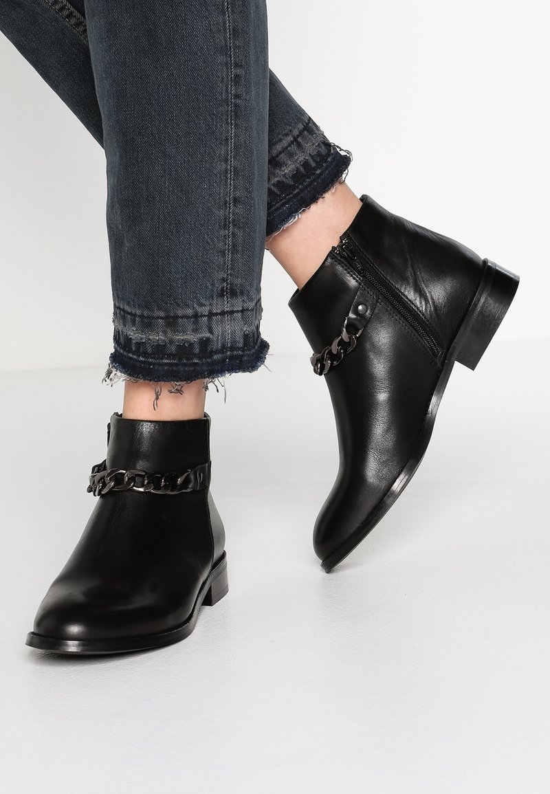 Botas de cuero negras hasta el tobillo con cremallera lateral y cadena de metal decorativa, combinadas con vaqueros oscuros de mezclilla con dobladillo deshilachado.