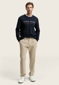 Camisa de manga larga azul marino con ribetes blancos, con texto rojo, combinada con pantalones beige ajustados y zapatillas blancas, sobre un fondo claro.