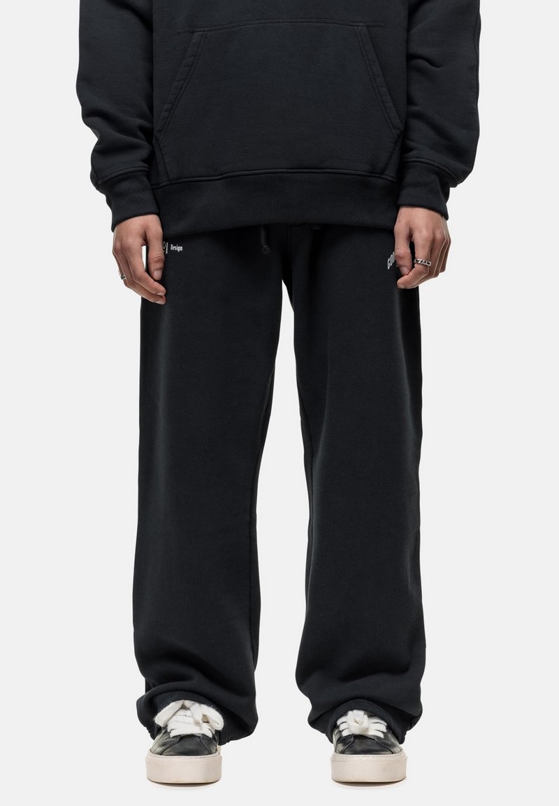 Zwarte katoenen hoodie met een voorzak, gecombineerd met losse zwarte sweatpants. Bevat witte logo-accenten en grijze sneakers met witte veters.