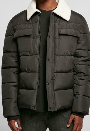 Donkergroene pufferjack met een witte fleece kraag, met twee borstzakken en een ritssluiting aan de voorkant, gedragen over een zwart shirt.